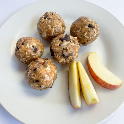 five peanut butter energy bites with three pieces of apple on a white plate.