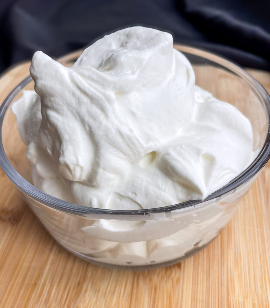 A glass bowl of homemade whipped cream on top of a wood cutting board