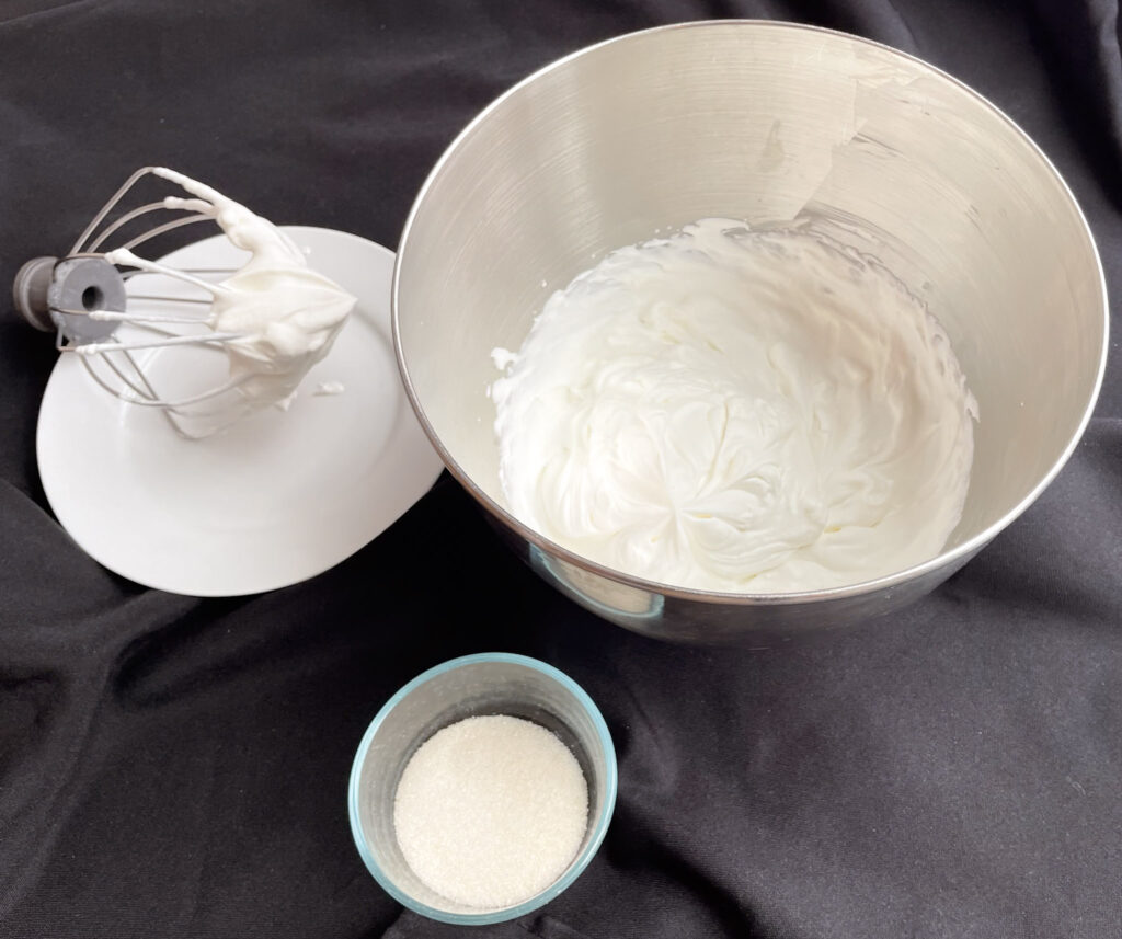 a metal whip and a metal bowl of whipped cream beside a small glass bowl of sugar